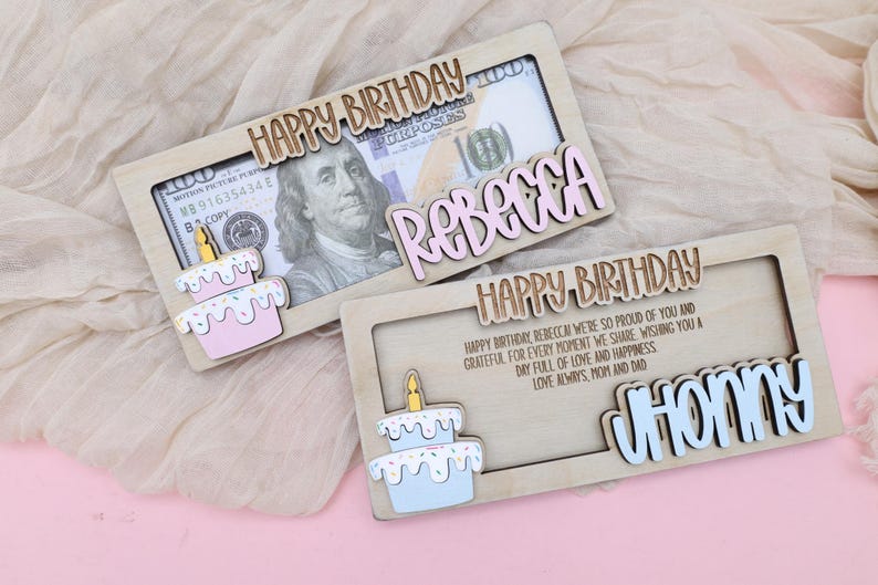 Puede incluir: Dos soportes de tarjetas de cumplea&ntilde;os de madera con una ranura para dinero en efectivo. La tarjeta superior tiene un pastel rosa y blanco con una vela amarilla y el texto "HAPPY BIRTHDAY REBECCA". La tarjeta inferior tiene un pastel azul y blanco con una vela amarilla y el texto "HAPPY BIRTHDAY CHONMY".