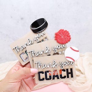 May include: Wooden thank you cards with sports-themed toppers. One card has a hockey puck, another a red pom-pom, and the last a baseball. The cards say "Thank you COACH" and "Thank you" in white script.