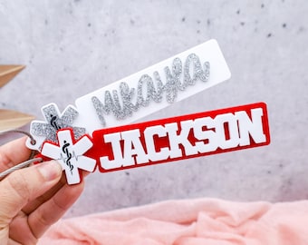 May include: Two personalized name tags are displayed. One is white with the name "Mikayla" in silver glitter. The other is red with the name "JACKSON" in white block letters. Both tags have a medical symbol.