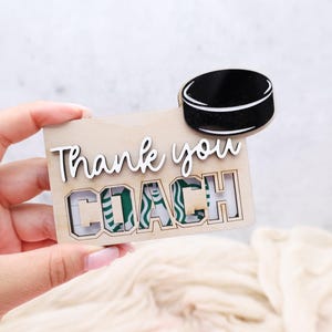 May include: A wooden gift card holder with the words "Thank you COACH" in white script. A black hockey puck is attached to the top. The gift card holder has a cutout for a Starbucks gift card.