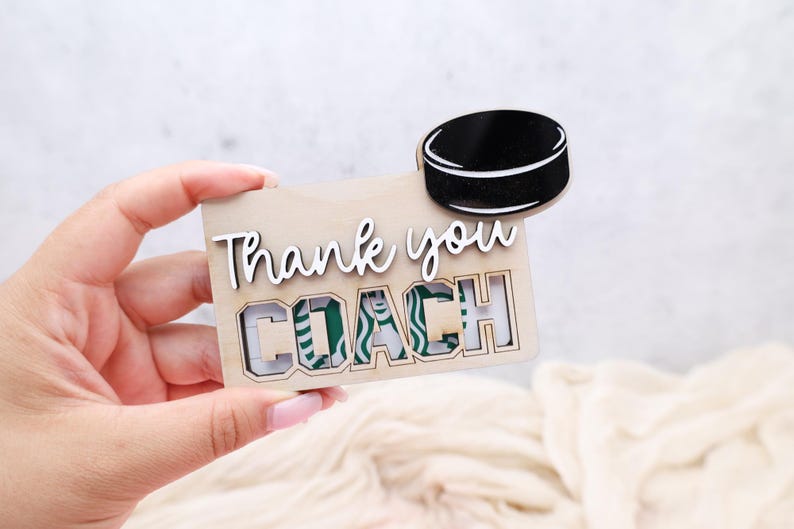 May include: A wooden gift card holder with the words "Thank you COACH" in white script. A black hockey puck is attached to the top. The gift card holder has a cutout for a Starbucks gift card.