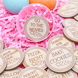 May include: Wooden activity tokens with various engraved phrases like "Go to the Movies" and "Bake Cookies." The tokens are scattered on pink shredded paper, with a pink bowl and colorful Easter eggs in the background. The tokens have a carrot illustration.