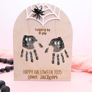 May include: Wooden Halloween decoration with handprint spiders. The sign has a white spider web and a black spider at the top. The text reads "Dropping by to say" and "Happy Halloween 2025 Love, Jackson".