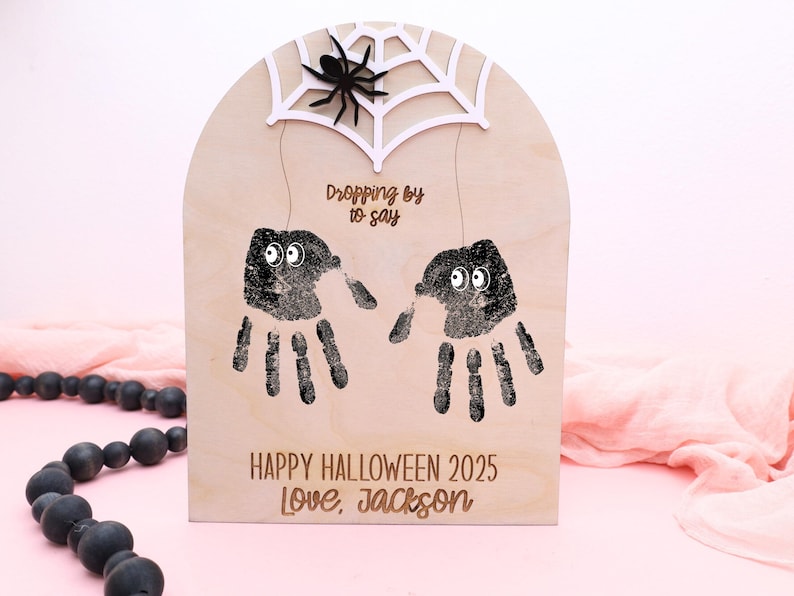 May include: Wooden Halloween decoration with handprint spiders. The sign has a white spider web and a black spider at the top. The text reads "Dropping by to say" and "Happy Halloween 2025 Love, Jackson".