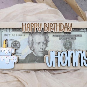 Puede incluir: Un marco de madera con un recorte de un pastel de cumplea&ntilde;os y las palabras "Feliz cumplea&ntilde;os" y "Johnny". Un billete de veinte euros es visible en el marco.