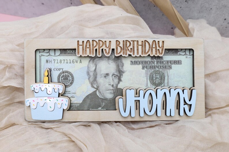 Puede incluir: Un marco de madera con un recorte de un pastel de cumplea&ntilde;os y las palabras "Feliz cumplea&ntilde;os" y "Johnny". Un billete de veinte euros es visible en el marco.