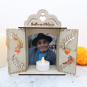 May include: A wooden memorial box with a photo of a man inside. The box has decorative cutouts, a heart, and the name "Guillermo Melgaza" at the top. A small, white LED candle sits in front of the photo.