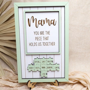 Puede incluir: Un marco de madera verde claro con fondo blanco y un mensaje que dice "Mama You are the piece that holds us together". El marco tiene un dise&ntilde;o de pieza de rompecabezas con nombres en cada pieza, incluyendo "Mama", "Crystal", "Amy", "Malcolm", "Noah", "Ricky", "Brandon", "Liam", y "Lisa".