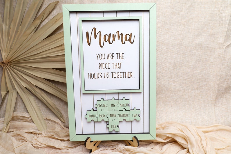 Puede incluir: Un marco de madera verde claro con fondo blanco y un mensaje que dice "Mama You are the piece that holds us together". El marco tiene un dise&ntilde;o de pieza de rompecabezas con nombres en cada pieza, incluyendo "Mama", "Crystal", "Amy", "Malcolm", "Noah", "Ricky", "Brandon", "Liam", y "Lisa".
