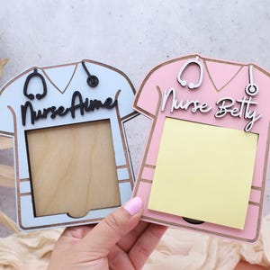 May include: Two nurse-themed memo holders shaped like scrubs tops. One is light blue with "Nurse Aimee" and the other is pink with "Nurse Betty". Each has a stethoscope and a wooden frame for notes.