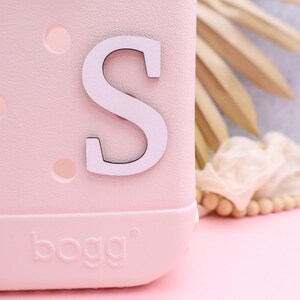 Pode incluir: Uma bolsa de pl&aacute;stico rosa com uma letra "S" branca presa na frente. A bolsa tem uma superf&iacute;cie texturizada e a palavra "bogg" est&aacute; impressa na parte inferior.