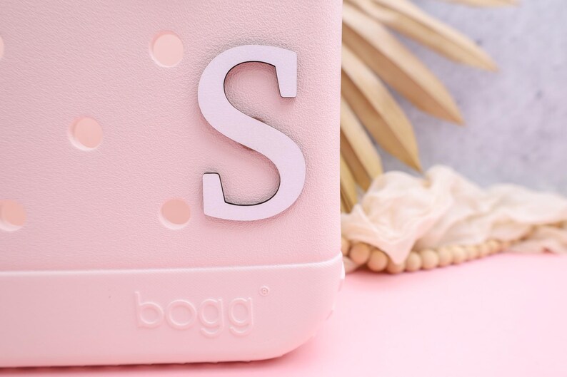 Pode incluir: Uma bolsa de pl&aacute;stico rosa com uma letra "S" branca presa na frente. A bolsa tem uma superf&iacute;cie texturizada e a palavra "bogg" est&aacute; impressa na parte inferior.