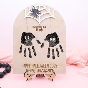 May include: Wooden Halloween sign with handprint spiders, a spider web, and a spider. The sign says "Dropping by to say" and "Happy Halloween 2025 Love, Jackson". The sign is on a wooden stand.