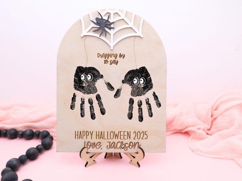 May include: Wooden Halloween sign with handprint spiders, a spider web, and a spider. The sign says "Dropping by to say" and "Happy Halloween 2025 Love, Jackson". The sign is on a wooden stand.