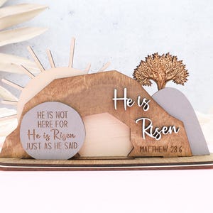 May include: Wooden Easter decoration with a brown, gray, and beige color scheme. The decoration features a sun, a tree, and a tomb with the text "He is not here for He is Risen just as He said" and "He is Risen Matthew 28:6".