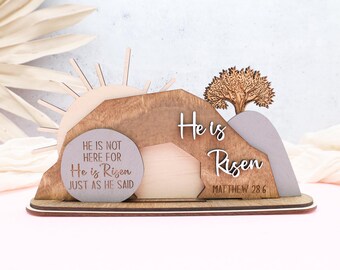 May include: Wooden Easter decoration with a brown, gray, and beige color scheme. The decoration features a sun, a tree, and a tomb with the text "He is not here for He is Risen just as He said" and "He is Risen Matthew 28:6".