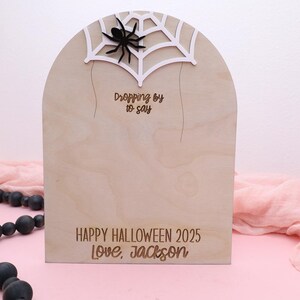 May include: Wooden Halloween decoration in the shape of a tombstone, featuring a spider and web design at the top. The text "Dropping by to say" is above "HAPPY HALLOWEEN 2025 Love, Jackson".