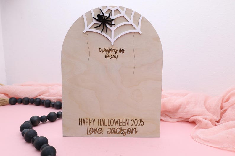 May include: Wooden Halloween decoration in the shape of a tombstone, featuring a spider and web design at the top. The text "Dropping by to say" is above "HAPPY HALLOWEEN 2025 Love, Jackson".