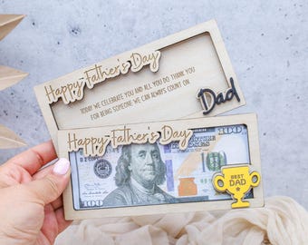 May include: Two wooden Father's Day gift card holders. The top one says "Happy Father's Day" and "Dad". The bottom one holds a $100 bill and has a yellow trophy that says "Best Dad".