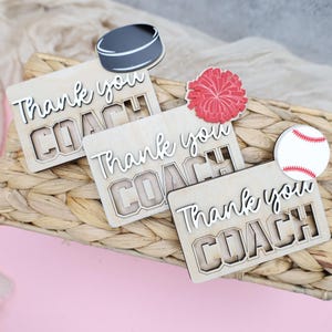 May include: Wooden plaques with the words "Thank you COACH" are displayed on a woven basket. A hockey puck, a red pom-pom, and a baseball are placed on top. The background is a light pink surface with a beige fabric.