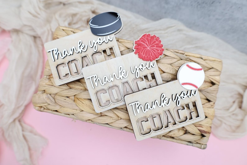 May include: Wooden plaques with the words "Thank you COACH" are displayed on a woven basket. A hockey puck, a red pom-pom, and a baseball are placed on top. The background is a light pink surface with a beige fabric.
