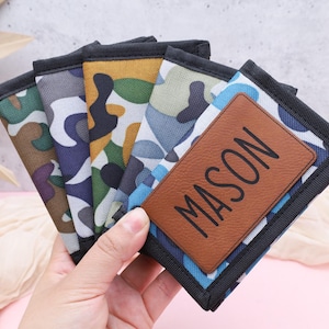 May include: A set of five camouflage patterned wallets with a brown leather patch that says "MASON" on the front.