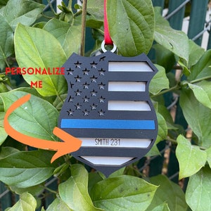 May include: A black and white wooden ornament shaped like a police badge with a thin blue line and an American flag design. The ornament has the text "SMITH 231" on the bottom.