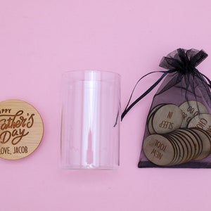 May include: A glass jar with a wooden lid that says "Happy Father's Day Love, Jacob". A black mesh bag filled with small wooden circles with words like "Sleep In" and "New Tool" is next to the jar.
