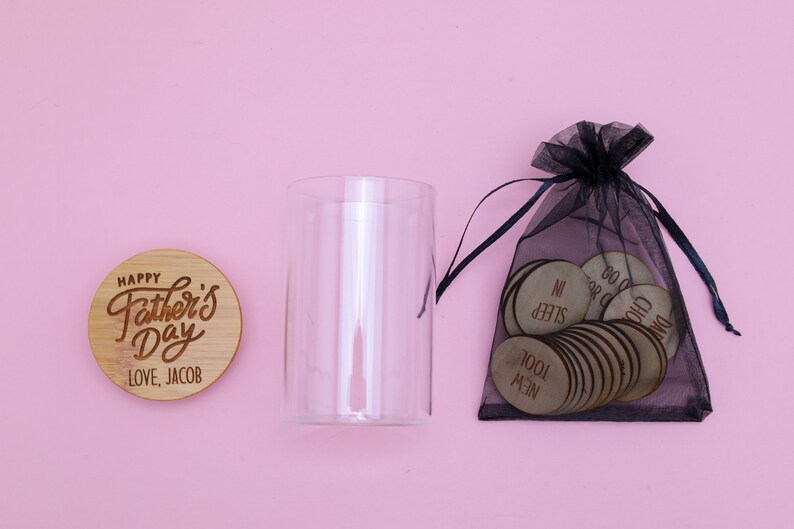 May include: A glass jar with a wooden lid that says "Happy Father's Day Love, Jacob". A black mesh bag filled with small wooden circles with words like "Sleep In" and "New Tool" is next to the jar.