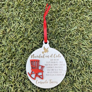 May include: White wooden Christmas ornament with a red rocking chair and gold text that reads "Navidad en el Cielo" and "Consuelo Torres". The text says "Que hacen? Ellos bajan del cielo para pasar tiempo contigo. Guardales un asiento, solo una silla vacia. No los podras ver, pero ellos estaran ahi!"