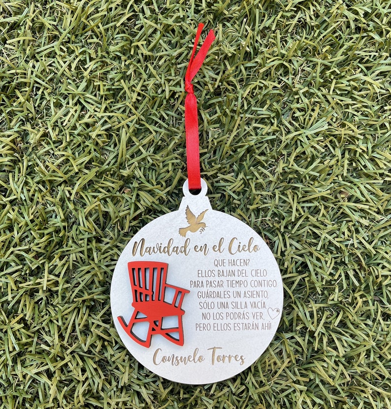 May include: White wooden Christmas ornament with a red rocking chair and gold text that reads "Navidad en el Cielo" and "Consuelo Torres". The text says "Que hacen? Ellos bajan del cielo para pasar tiempo contigo. Guardales un asiento, solo una silla vacia. No los podras ver, pero ellos estaran ahi!"
