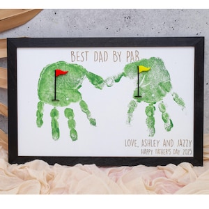 May include: A framed print with green handprints forming a golf-themed design. The text "BEST DAD BY PAR" is at the top, with "LOVE, ASHLEY AND JAZZY HAPPY FATHER'S DAY 2025" at the bottom. Red and yellow flags are included.