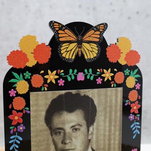 May include: A black acrylic picture frame featuring a monarch butterfly at the top center. The frame is decorated with colorful printed marigold flowers, small blossoms, and green leaves. A black and white portrait of a person is inside the frame.