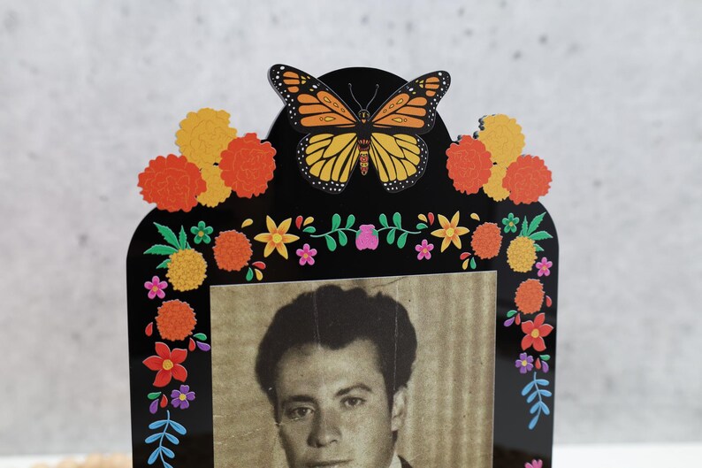 May include: A black acrylic picture frame featuring a monarch butterfly at the top center. The frame is decorated with colorful printed marigold flowers, small blossoms, and green leaves. A black and white portrait of a person is inside the frame.