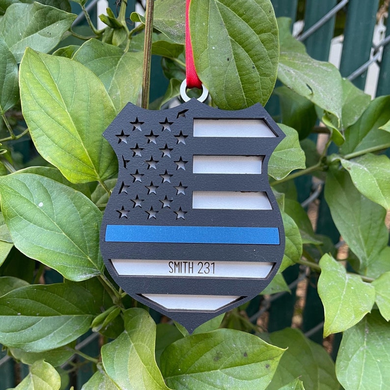 May include: A black wooden ornament shaped like a police badge with a thin blue line and an American flag design. The ornament has the text "SMITH 231" on it.
