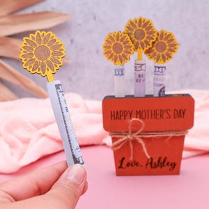 Op de afbeelding: Een houten plantenbak met de woorden "Happy Mother's Day" en "Love, Ashley" erop gegraveerd. De plantenbak is versierd met vier gele houten zonnebloemen, die elk een opgerold dollarbiljet vasthouden.