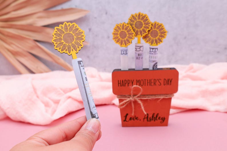 Op de afbeelding: Een houten plantenbak met de woorden "Happy Mother's Day" en "Love, Ashley" erop gegraveerd. De plantenbak is versierd met vier gele houten zonnebloemen, die elk een opgerold dollarbiljet vasthouden.