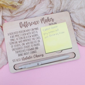 May include: Wooden desk organizer with the text "Difference Maker" and a definition. Includes a yellow sticky note with handwritten notes and a pen. The organizer is on a pink surface.