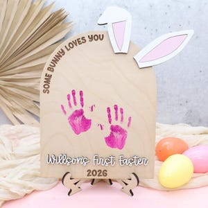 Puede incluir: Cartel de madera con temática de Pascua con orejas de conejo y el texto "Some Bunny Loves You". Presenta huellas de manos rosas y la inscripción "Willson's first Easter 2026". Incluye un soporte y huevos de Pascua decorativos.