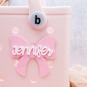 May include: A pink tote bag with a white bow and the name "Jennifer" on it. The bag has a white button with the letter "b" on it.