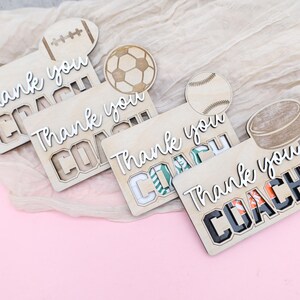 May include: Wooden sports-themed thank you cards with the words "Thank you COACH" in white script. Each card features a different sport: football, soccer, baseball, and hockey. The cards are on a light pink surface.
