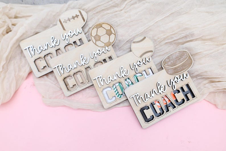 May include: Wooden sports-themed thank you cards with the words "Thank you COACH" in white script. Each card features a different sport: football, soccer, baseball, and hockey. The cards are on a light pink surface.