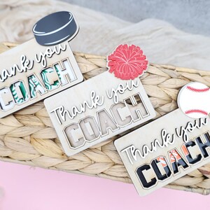 May include: Wooden plaques with the words "Thank you COACH" in a cutout design. Each plaque has a different sports-themed accent: a hockey puck, a red pom-pom, and a baseball. The plaques are on a woven basket.