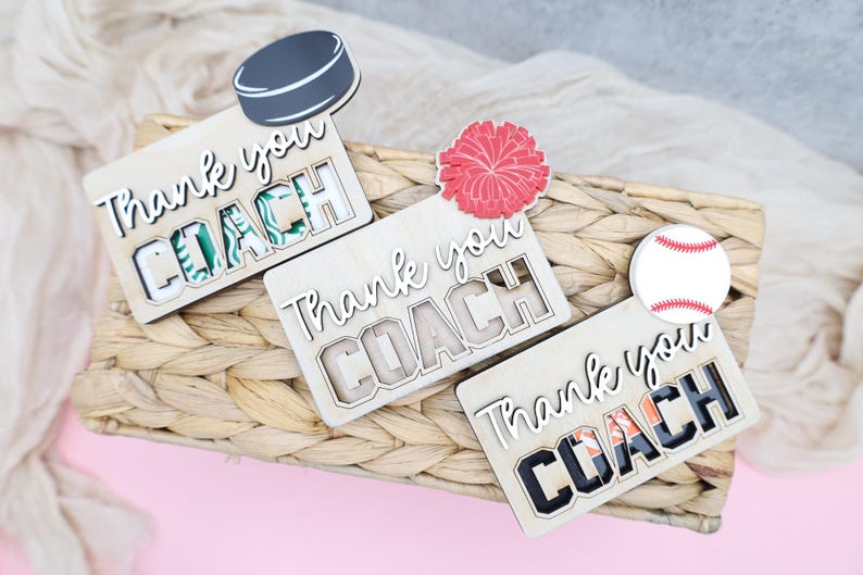 May include: Wooden plaques with the words "Thank you COACH" in a cutout design. Each plaque has a different sports-themed accent: a hockey puck, a red pom-pom, and a baseball. The plaques are on a woven basket.
