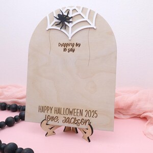 May include: A wooden Halloween sign with a spider web and spider design. The sign reads "Dropping by to say" and "Happy Halloween 2025 Love, Jackson". The sign is on a wooden stand.