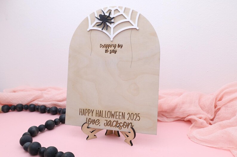 May include: A wooden Halloween sign with a spider web and spider design. The sign reads "Dropping by to say" and "Happy Halloween 2025 Love, Jackson". The sign is on a wooden stand.