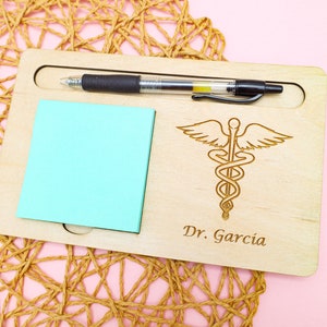 May include: A wooden desk organizer with a pen holder, a sticky note pad, and a laser engraved medical symbol with the text "Dr. Garcia".