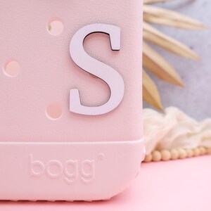 Pode incluir: Uma bolsa Bogg rosa com uma letra S branca na frente.