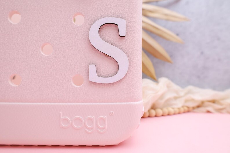 Pode incluir: Uma bolsa Bogg rosa com uma letra S branca na frente.