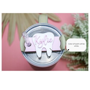 May include: A pink and white tooth-shaped tumbler lid with the name "Kylie" written on it. The tooth is white and the name is pink.